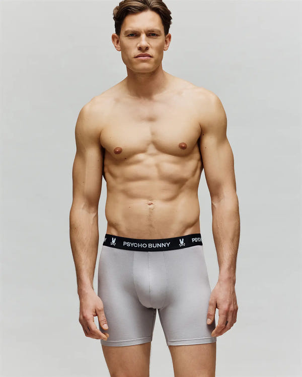 A muscular man stands against a plain background, wearing Psycho Bunny MENS 3 PACK BOXER BRIEF - MO1400006 boxer briefs with a black waistband that reads "PSYCHO BUNNY" in white. He faces forward with a neutral expression.