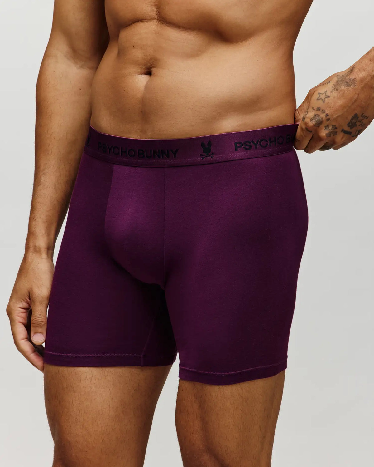 A person wears deep purple Psycho Bunny MENS 3 PACK BOXER BRIEF - MO1400008 with a black waistband showing the brand name and bunny logo. Torso and upper thighs are visible against a plain, light background.