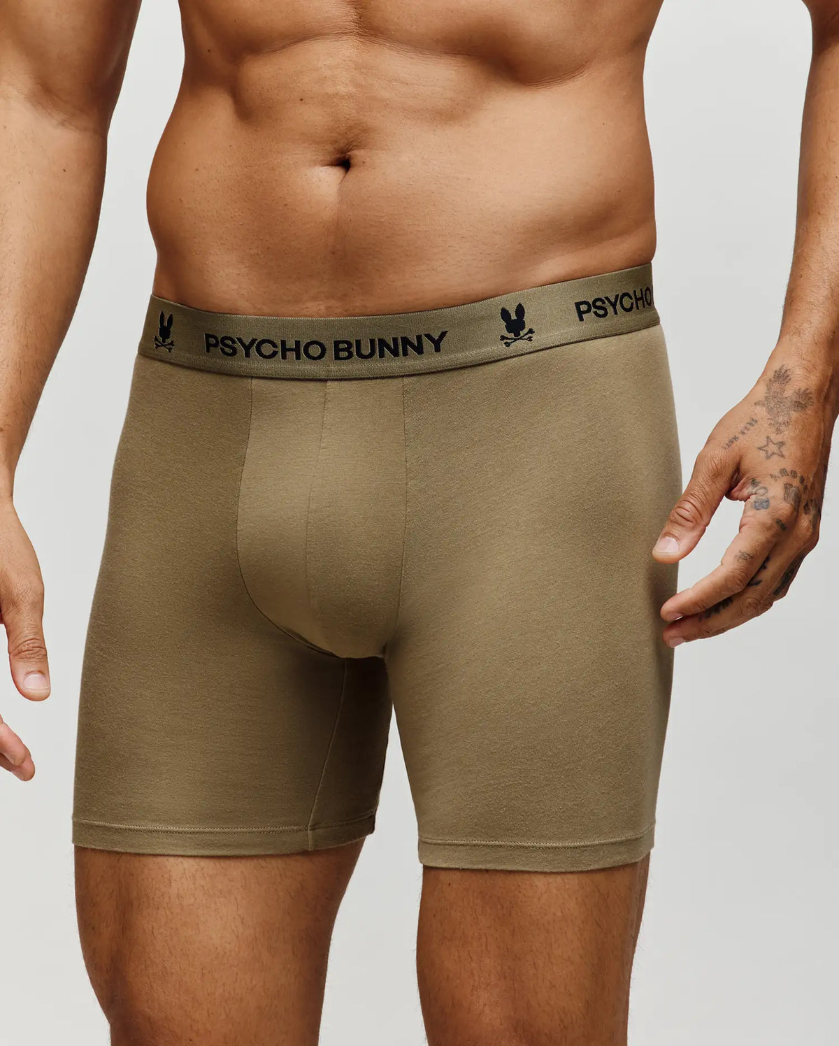 A man wearing Psycho Bunny MENS 3 PACK BOXER BRIEF - MO1400008 in olive green cotton modal, featuring a black Psycho Bunny logo waistband; his torso and tattooed hand are partially visible.