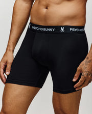 A person wears black Psycho Bunny MENS 3 PACK BOXER BRIEF - MO1400008 against a plain background, hands by their sides. The waistband displays the Psycho Bunny logo and text, showcasing stylish men's underwear.