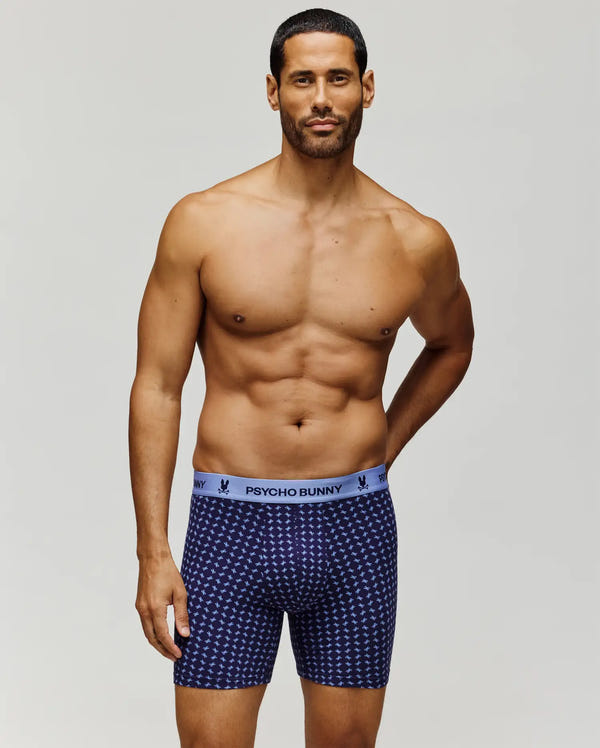 A fit, bearded man models Psycho Bunny’s MENS SINGLE BOXER BRIEF - MO1400010, featuring breathable blue Pima cotton with a white pattern and light blue waistband. He stands topless against a plain background, looking confidently at the camera.