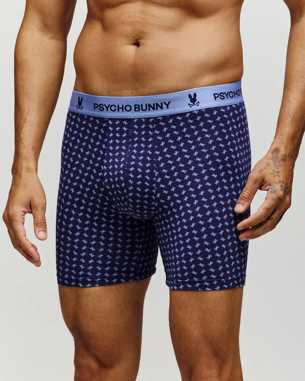A man wears Psycho Bunny's MENS SINGLE BOXER BRIEF (MO1400010), featuring navy blue fabric with light blue bunny patterns, a light blue logo waistband, and breathable Pima cotton knit for comfort.