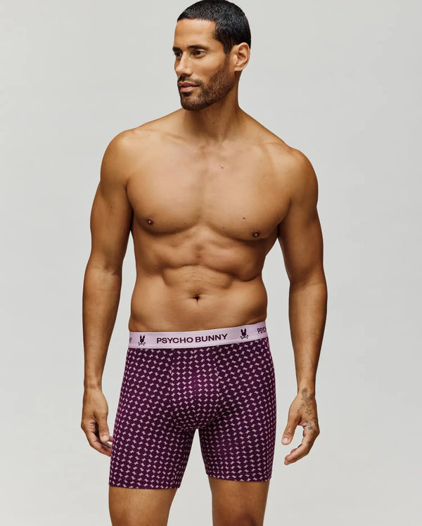 A man stands against a plain background wearing Psycho Bunny MENS SINGLE BOXER BRIEF - MO1400010 in maroon print, crafted from breathable knit Pima cotton with a light pink "PSYCHO BUNNY" waistband for all-day comfort.