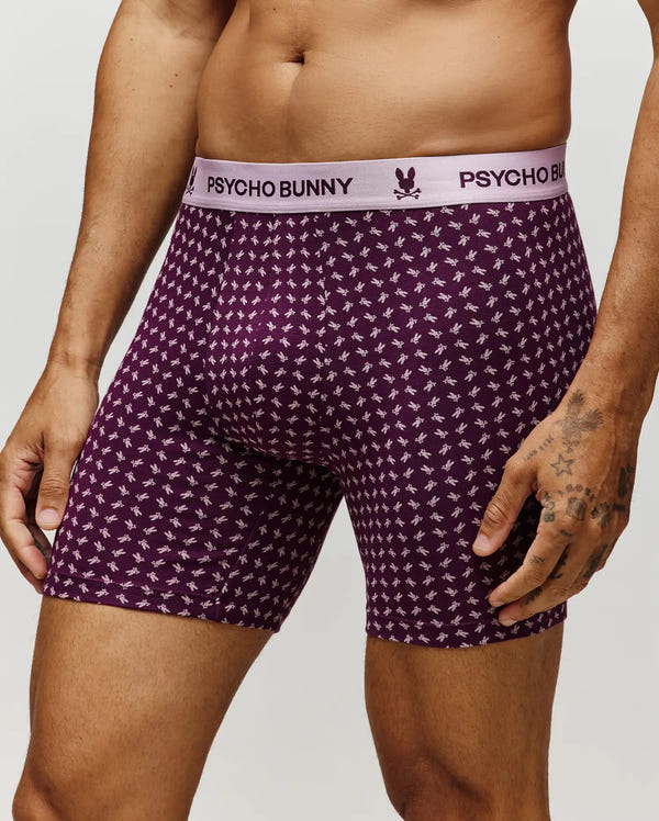 A person wears Psycho Bunny’s MENS SINGLE BOXER BRIEF - MO1400010: purple, breathable Pima cotton, small white bunny print, and a white waistband with “Psycho Bunny” in purple. Only the lower torso and upper legs are shown.
