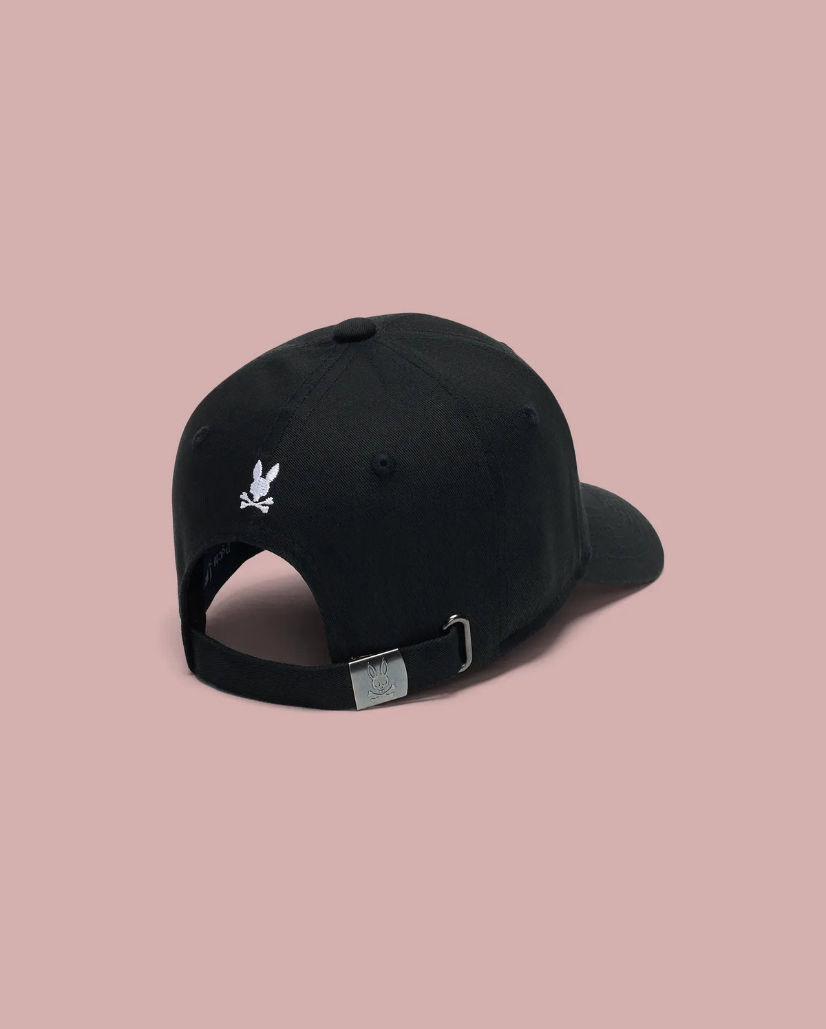 The MENS ARCHIVE BASEBALL CAP (MO1500002) by Psycho Bunny is shown from the back in black cotton, featuring a small white embroidered Bunny logo above its adjustable strap, set against a plain light pink background.