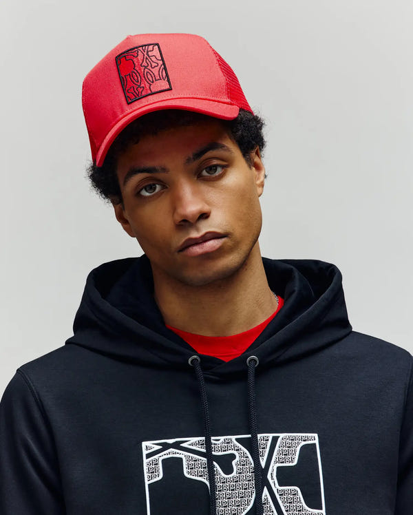 A young person wears the Psycho Bunny MENS GLENN TRUCKER CAP - MO1500004, an adjustable red snapback with a black abstract design, paired with a black hoodie and red shirt, standing against a plain light background.