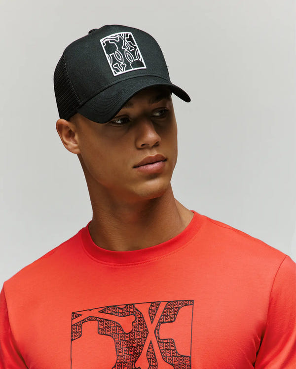 A young man in a Psycho Bunny MENS GLENN TRUCKER CAP (MO1500004) with a white logo and a bright red T-shirt featuring the matching Bunny graphic looks off to the side against a plain background.