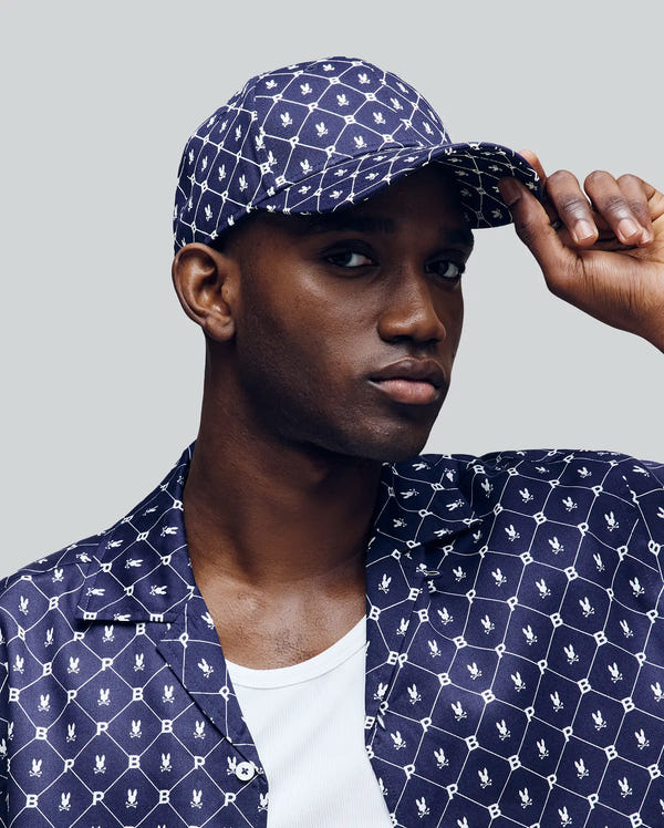 A person poses against a plain light grey background wearing the Psycho Bunny MENS HUNTER BASEBALL CAP - MO1500007 in navy blue with PB print and an adjustable strap, paired with a matching shirt featuring a white geometric bunny pattern and a white undershirt.