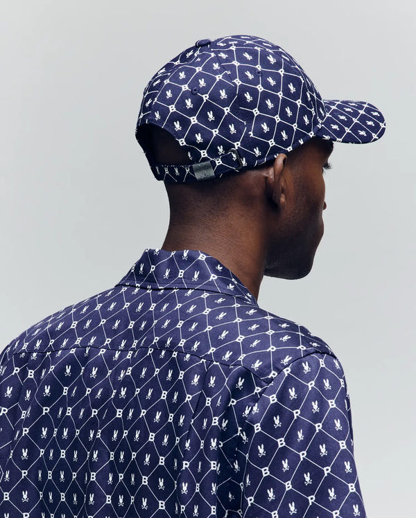 A person is seen from behind wearing a matching navy shirt and the Psycho Bunny MENS HUNTER BASEBALL CAP - MO1500007, both with a white geometric PB print featuring birds and "M" in diamonds. The cap has an adjustable strap for comfort.