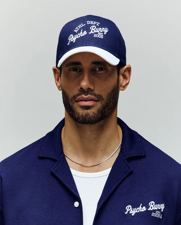 A bearded man wears a navy blue Psycho Bunny MENS MATTEO BASEBALL CAP (MO1500008) with an embroidered wordmark and a matching collared shirt over a white t-shirt, standing against a plain light background.