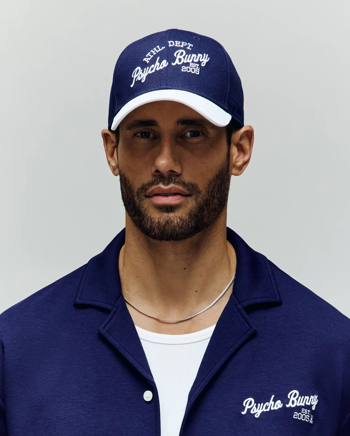 A bearded man wears a navy blue Psycho Bunny MENS MATTEO BASEBALL CAP (MO1500008) with an embroidered wordmark and a matching collared shirt over a white t-shirt, standing against a plain light background.
