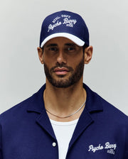 A bearded man wears a navy blue Psycho Bunny MENS MATTEO BASEBALL CAP (MO1500008) with an embroidered wordmark and a matching collared shirt over a white t-shirt, standing against a plain light background.