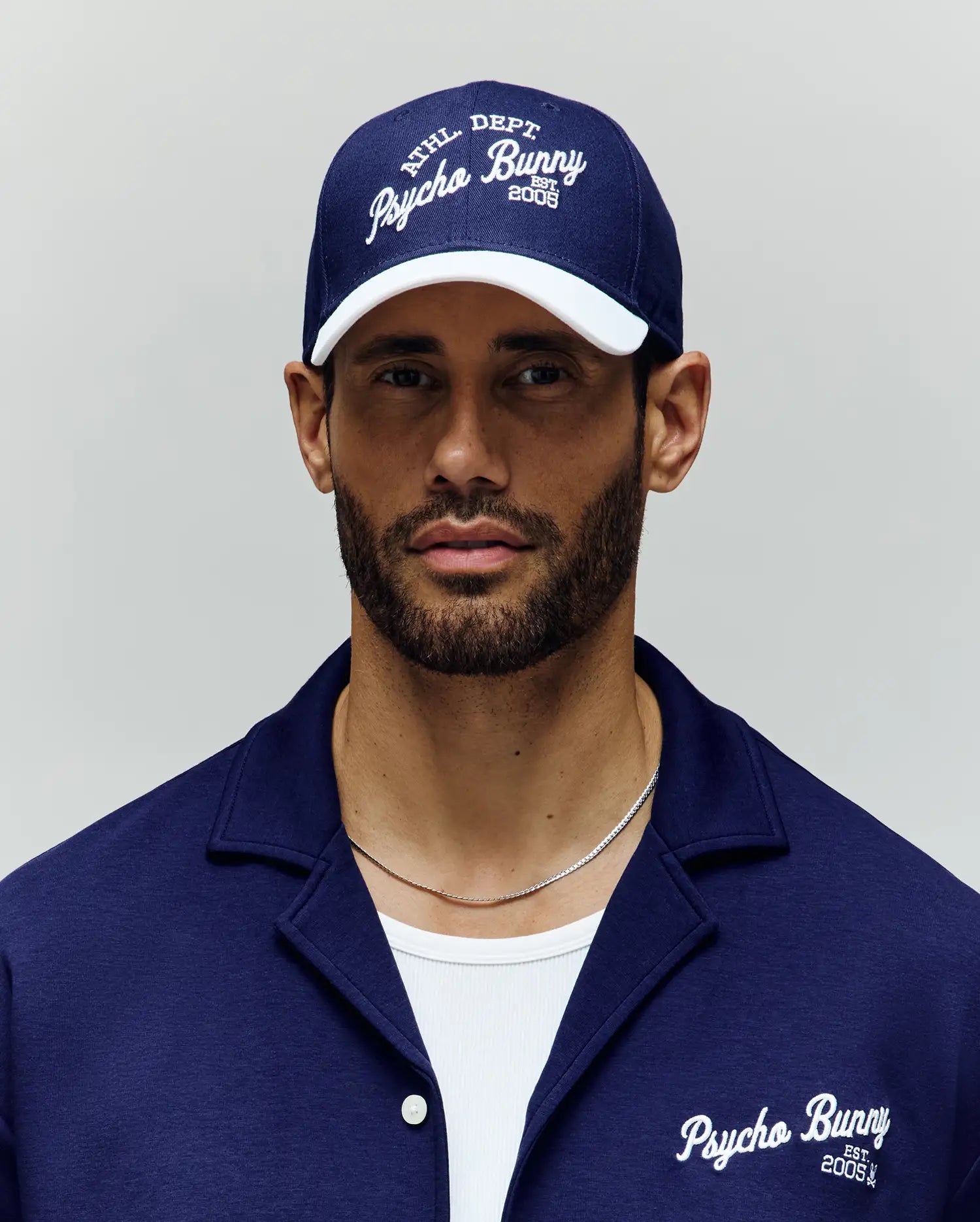 A bearded man wears a navy blue Psycho Bunny MENS MATTEO BASEBALL CAP (MO1500008) with an embroidered wordmark and a matching collared shirt over a white t-shirt, standing against a plain light background.