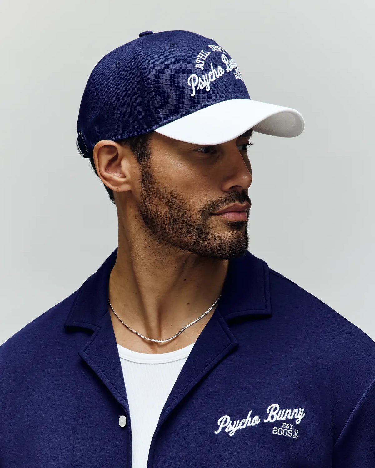A bearded man looks to the side, wearing the Psycho Bunny MENS MATTEO BASEBALL CAP - MO1500008 in navy and white, paired with a matching varsity shirt featuring an embroidered logo, and a white 100% cotton T-shirt underneath.
