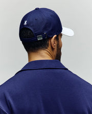 A man is seen from behind wearing a navy blue, 100% cotton shirt and the Psycho Bunny MENS MATTEO BASEBALL CAP (MO1500008), a varsity-style navy cap with white brim, embroidered wordmark logo, and silver buckle on the strap.