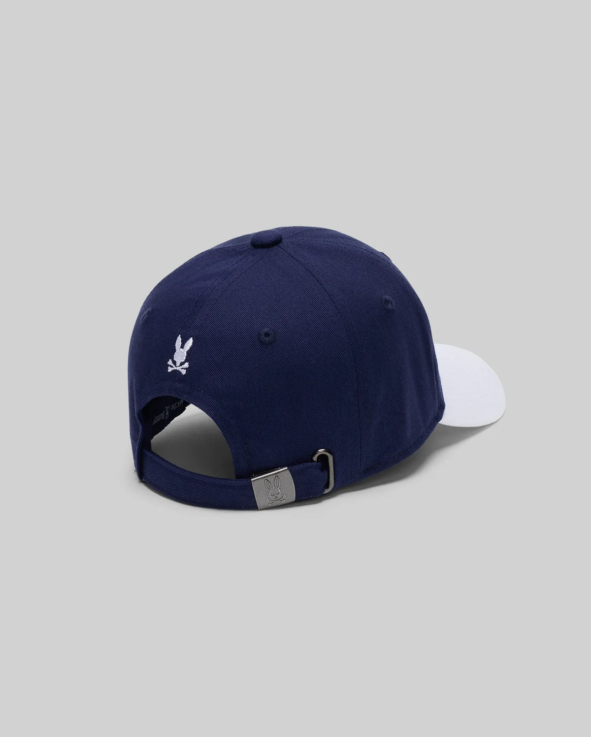 The MENS MATTEO BASEBALL CAP (MO1500008) by Psycho Bunny is a navy varsity-style cap with a white brim, small white embroidered wordmark logo, and adjustable metal-buckle strap, shown on a light gray background.