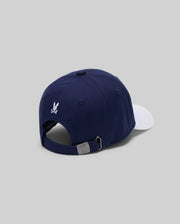 The MENS MATTEO BASEBALL CAP (MO1500008) by Psycho Bunny is a navy varsity-style cap with a white brim, small white embroidered wordmark logo, and adjustable metal-buckle strap, shown on a light gray background.