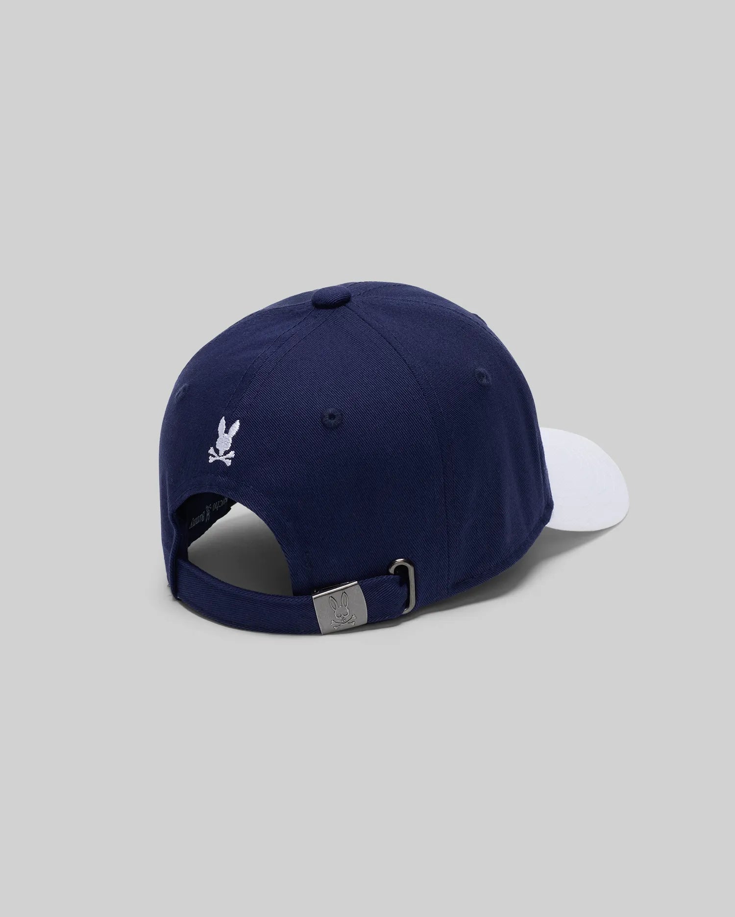 The MENS MATTEO BASEBALL CAP (MO1500008) by Psycho Bunny is a navy varsity-style cap with a white brim, small white embroidered wordmark logo, and adjustable metal-buckle strap, shown on a light gray background.