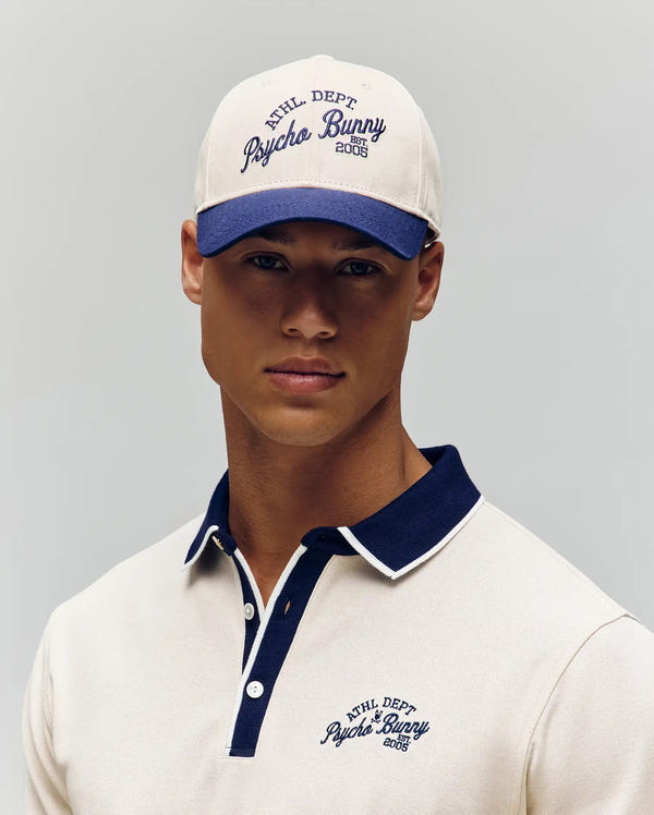 A man wears a cream Psycho Bunny MatteO Baseball Cap (MO1500008) with navy trim and "Psycho Bunny Athl Dept 2005" logo, featuring an adjustable strap, against a plain light background.