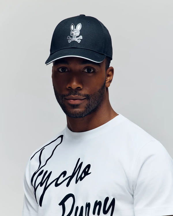 A man wears a Psycho Bunny MENS DEREK BASEBALL CAP - MO1500009 in black with a cartoon bunny and crossbones logo, paired with a white premium cotton T-shirt featuring black "Psycho Bunny" script, against a light-gray background.