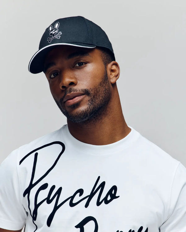 A man with a short beard wears the Psycho Bunny MENS DEREK BASEBALL CAP - MO1500009, featuring a bold white skull logo, and a white "Psycho Bunny" T-shirt. He faces the camera with a neutral expression against a plain background.