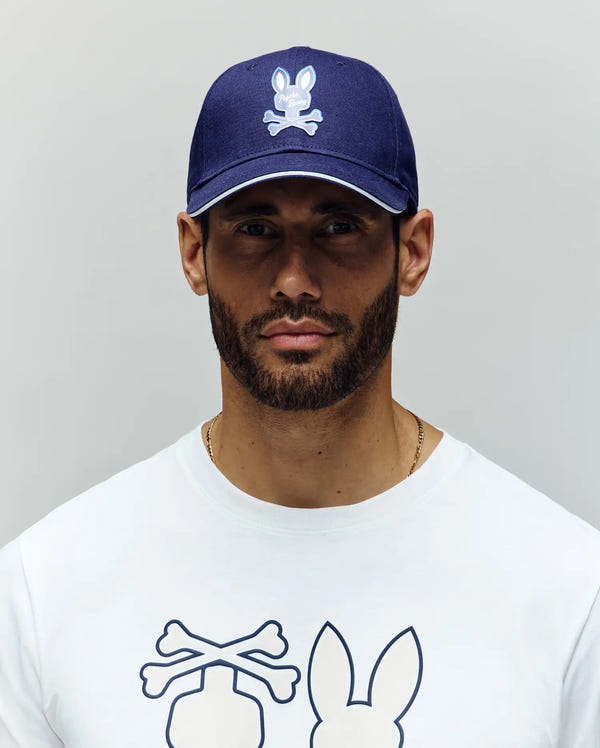 A man with a trimmed beard wears the Psycho Bunny MENS DEREK BASEBALL CAP – MO1500009 in blue, paired with a white premium cotton T-shirt, both featuring the bold bunny skull-and-crossbones logo, against a light gray backdrop.