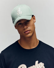 Wearing the Psycho Bunny MENS DEREK BASEBALL CAP - MO1500009 in light green with a bold bunny and crossbones logo, paired with a black premium cotton T-shirt featuring embroidered text, someone poses against a plain light background.