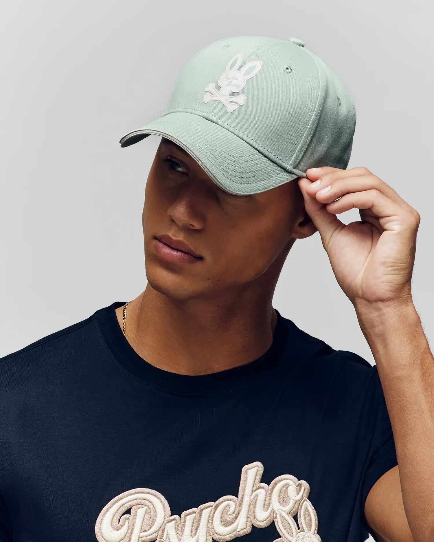 A person wearing the Psycho Bunny MENS DEREK BASEBALL CAP - MO1500009 in light green with a cartoon bunny and crossbones logo, and a black "Psycho" script T-shirt, tilts the cap while looking to the side.