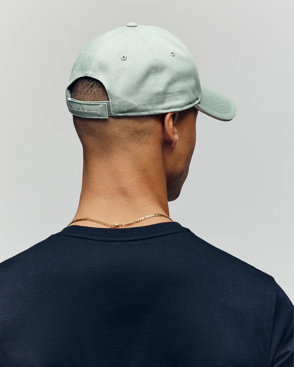 A person is shown from behind wearing the Psycho Bunny MENS DEREK BASEBALL CAP - MO1500009 in light green with a small tag above the adjustable strap, paired with a dark shirt and a gold chain necklace.