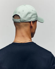 A person is shown from behind wearing the Psycho Bunny MENS DEREK BASEBALL CAP - MO1500009 in light green with a small tag above the adjustable strap, paired with a dark shirt and a gold chain necklace.