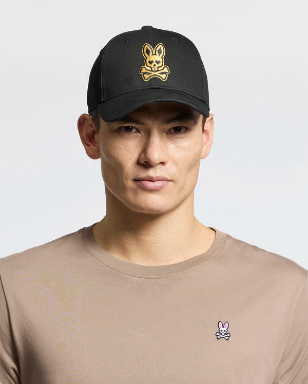 A person is wearing Psycho Bunny's MENS LAS VEGAS BASEBALL CAP (MO1500011) featuring a black cotton cap with a gold skull and crossbones bunny design and a beige shirt with intricate embroidery, including a small pink bunny logo on the chest, both made in China.