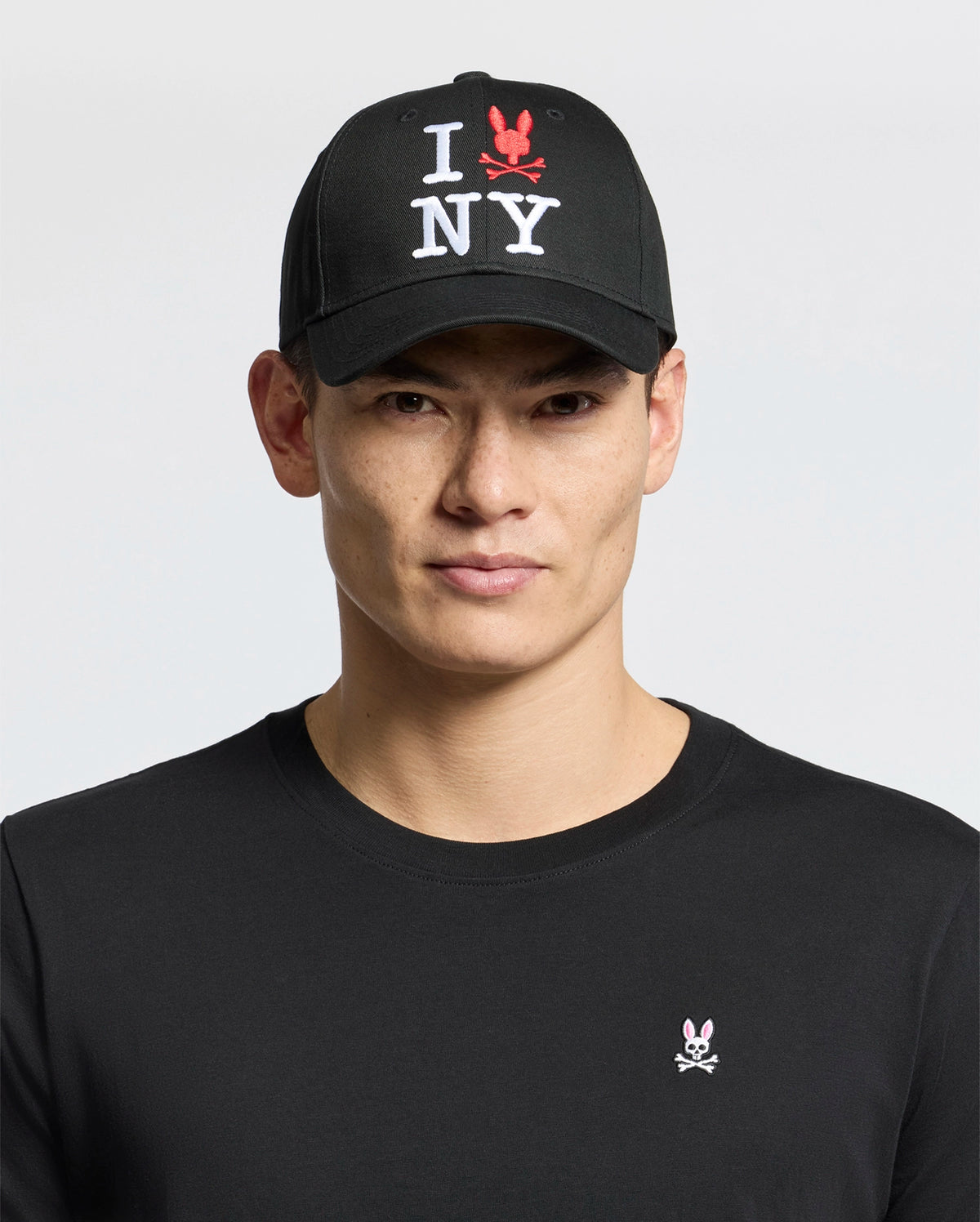 A man wears the Psycho Bunny MENS NEW YORK BASEBALL CAP - MO1500012, a black cotton cap with "I NY" and red embroidered bunny logo, plus a black shirt featuring a small pink and white embroidered bunny chest logo, against a light background.