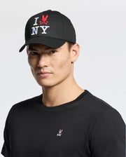 A man wearing a black cotton t-shirt and a Psycho Bunny MENS NEW YORK BASEBALL CAP (MO1500012) with "I ♥️ NY" embroidered in white and red stands against a plain background.