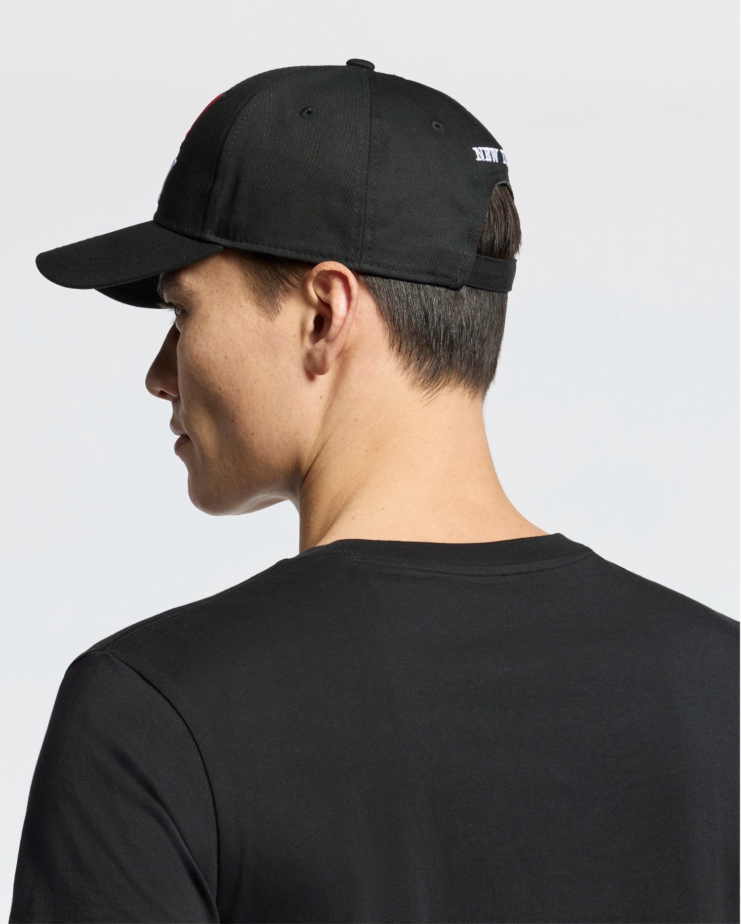 A person is shown from the back and side, facing left, wearing a Psycho Bunny MENS NEW YORK BASEBALL CAP - MO1500012 and a black t-shirt. The plain light background highlights the cap’s subtle embroidery.