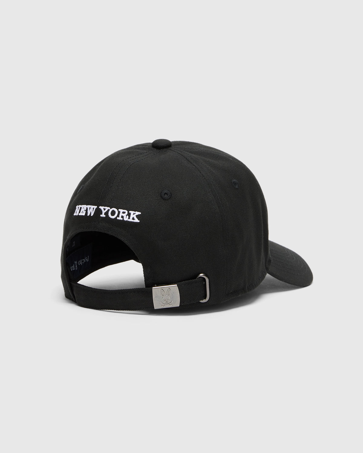 The Psycho Bunny MENS NEW YORK BASEBALL CAP (MO1500012) is a black woven cotton cap featuring "NEW YORK" embroidered in white above the adjustable strap with a small silver buckle at the back.