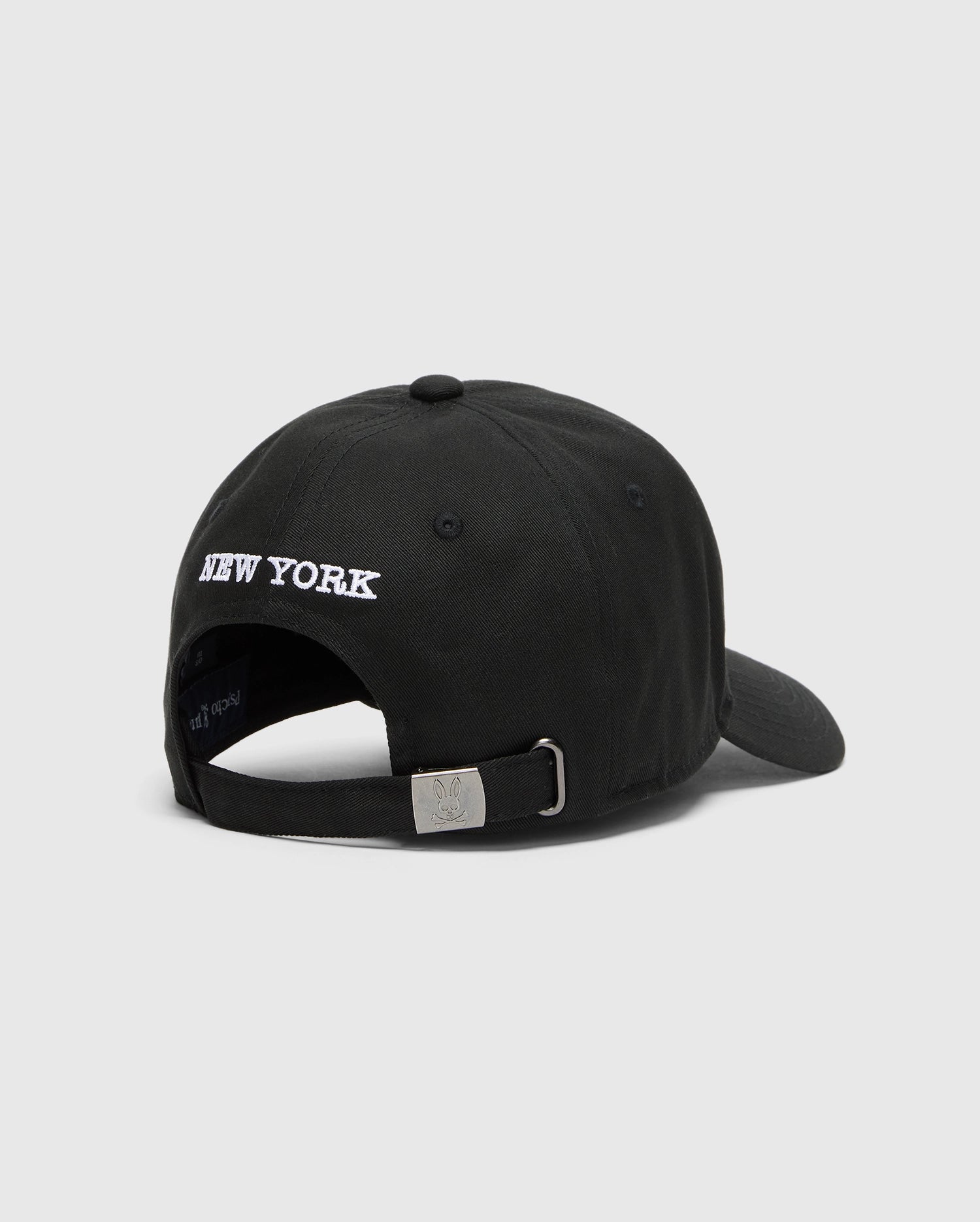 The Psycho Bunny MENS NEW YORK BASEBALL CAP (MO1500012) is a black woven cotton cap featuring "NEW YORK" embroidered in white above the adjustable strap with a small silver buckle at the back.