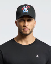 A man in a black Psycho Bunny MENS LOS ANGELES BASEBALL CAP features an embroidered bunny skull and palm trees logo. His soft cotton black shirt sports a small woven bunny skull emblem on the chest, set against a neutral background.