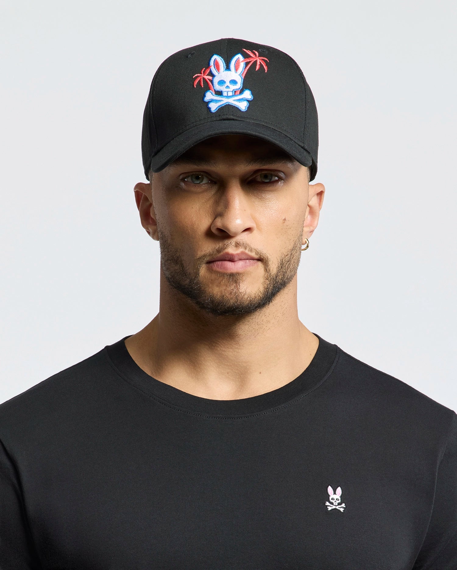 A man in a black Psycho Bunny MENS LOS ANGELES BASEBALL CAP features an embroidered bunny skull and palm trees logo. His soft cotton black shirt sports a small woven bunny skull emblem on the chest, set against a neutral background.