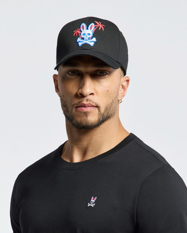 A person wearing the Psycho Bunny MENS LOS ANGELES BASEBALL CAP - MO1500013, which is black with an embroidered colorful skull and crossbones design featuring a rabbit and palm trees, pairs it with a black cotton shirt showcasing a small woven rabbit logo against a plain white background.