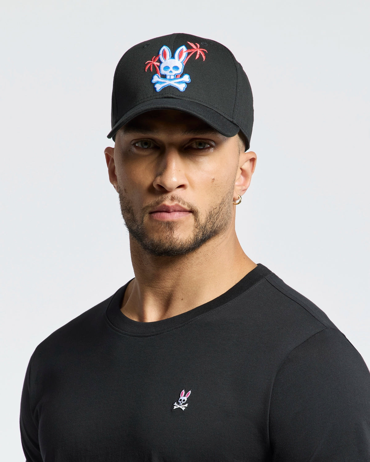 A person wearing the Psycho Bunny MENS LOS ANGELES BASEBALL CAP - MO1500013, which is black with an embroidered colorful skull and crossbones design featuring a rabbit and palm trees, pairs it with a black cotton shirt showcasing a small woven rabbit logo against a plain white background.