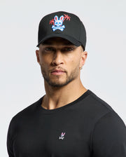 A person wearing the Psycho Bunny MENS LOS ANGELES BASEBALL CAP - MO1500013, which is black with an embroidered colorful skull and crossbones design featuring a rabbit and palm trees, pairs it with a black cotton shirt showcasing a small woven rabbit logo against a plain white background.