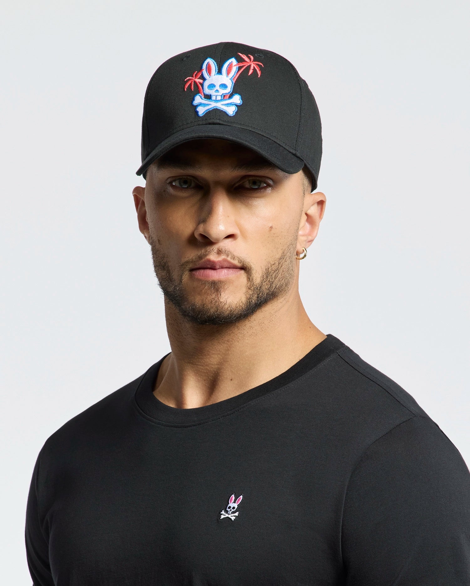 A person wearing the Psycho Bunny MENS LOS ANGELES BASEBALL CAP - MO1500013, which is black with an embroidered colorful skull and crossbones design featuring a rabbit and palm trees, pairs it with a black cotton shirt showcasing a small woven rabbit logo against a plain white background.
