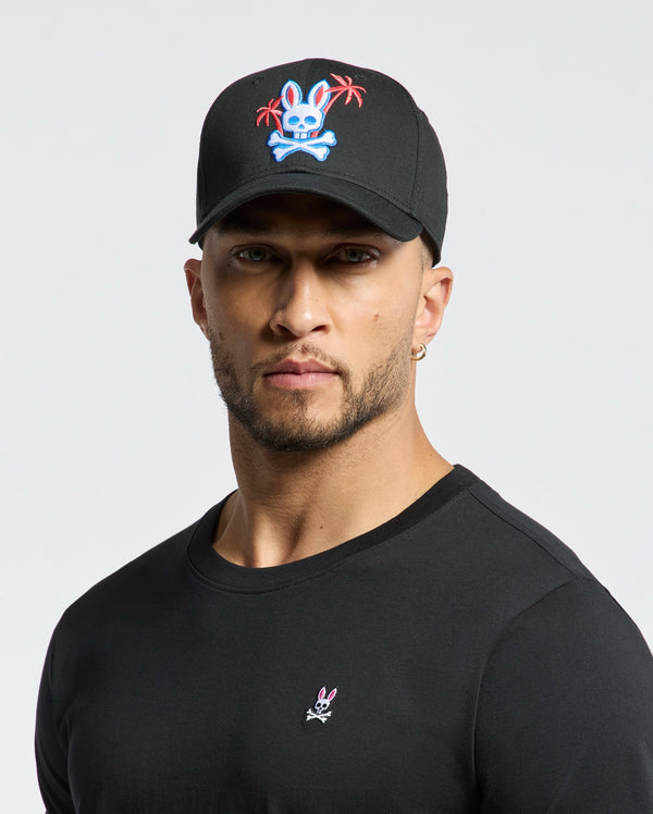 A person wearing the Psycho Bunny MENS LOS ANGELES BASEBALL CAP - MO1500013, which is black with an embroidered colorful skull and crossbones design featuring a rabbit and palm trees, pairs it with a black cotton shirt showcasing a small woven rabbit logo against a plain white background.