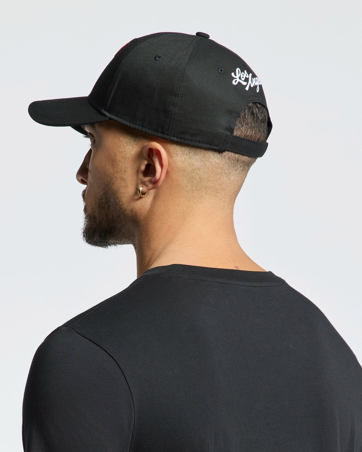 A person is seen from the back wearing a Psycho Bunny Los Angeles baseball cap (MO1500013) in black, featuring white embroidery. The cap is worn backward, displaying their short hair and earring. They're also wearing a black shirt against a plain white background.