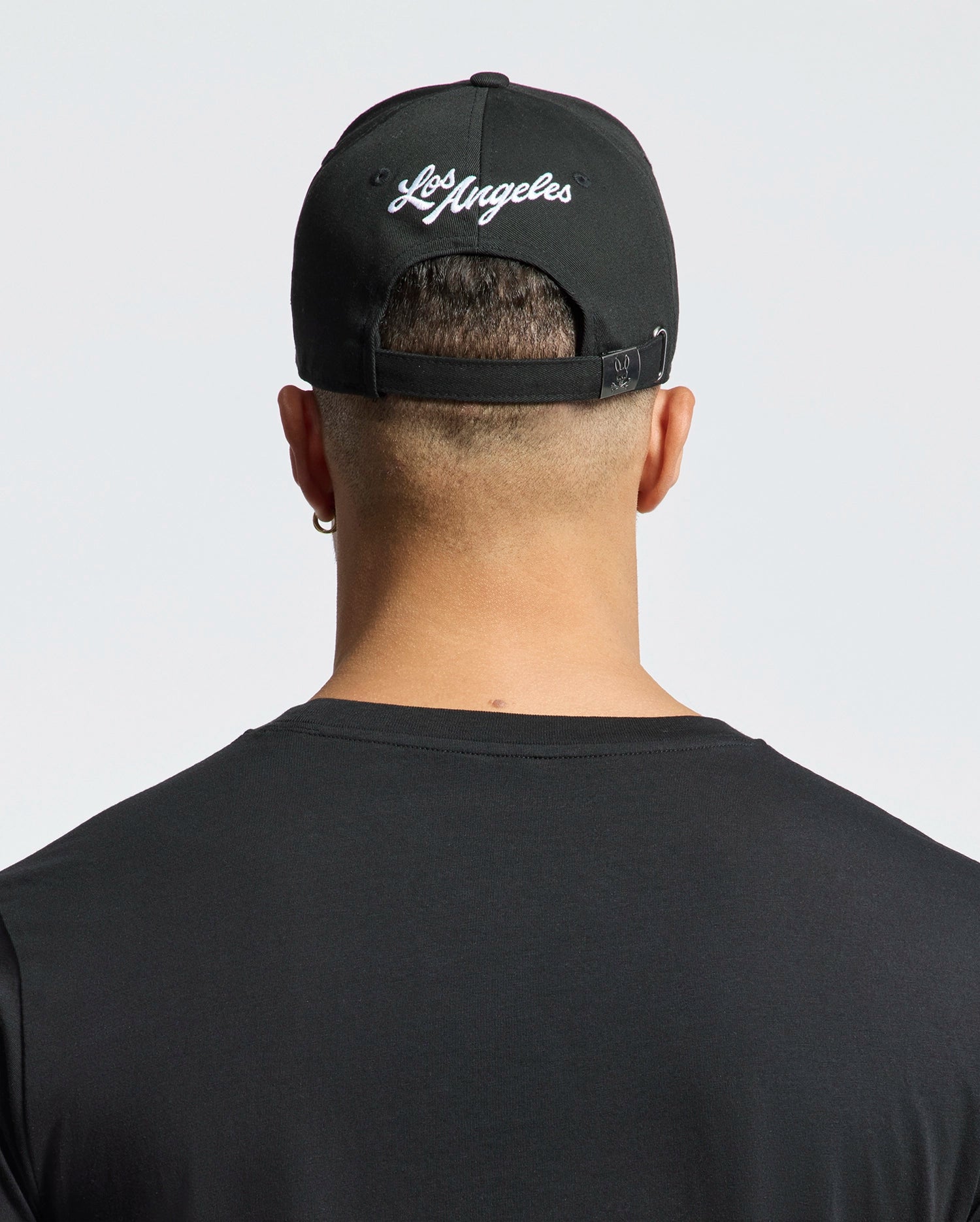 A person is shown from behind wearing a Psycho Bunny MENS LOS ANGELES BASEBALL CAP (MO1500013) with "Los Angeles" embroidered on the back, a black cotton t-shirt, and the cap's strap adjusted against a plain white background.