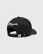 The Psycho Bunny MENS LOS ANGELES BASEBALL CAP (MO1500013) is a black cotton cap featuring "Los Angeles" embroidered in white on the back, with an adjustable strap, metal buckle, and a small woven logo.