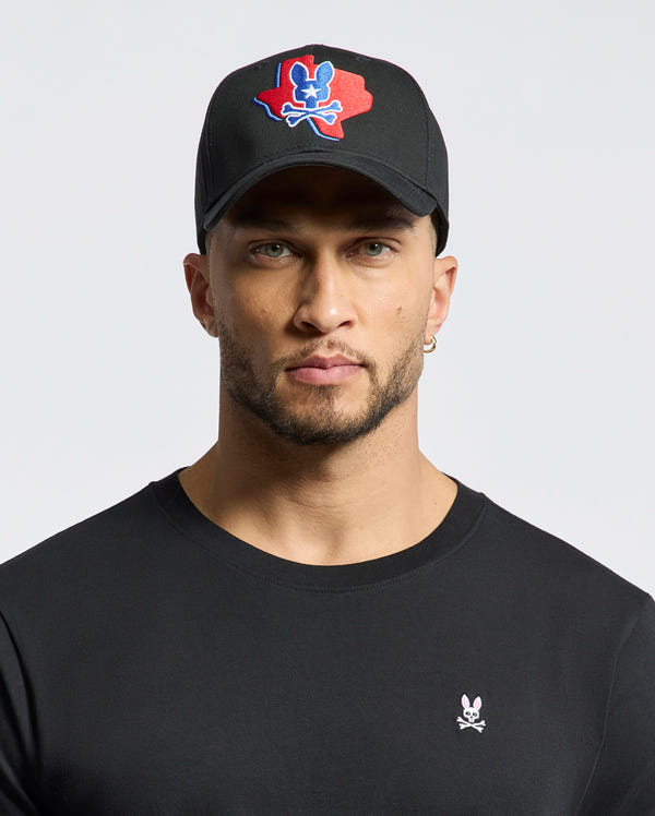 A man with light stubble wears a black Psycho Bunny MENS TEXAS BASEBALL CAP - MO1500014, featuring an embroidered red and blue skull design. He also sports a black shirt with a small woven skull and bunny logo against a plain white background.
