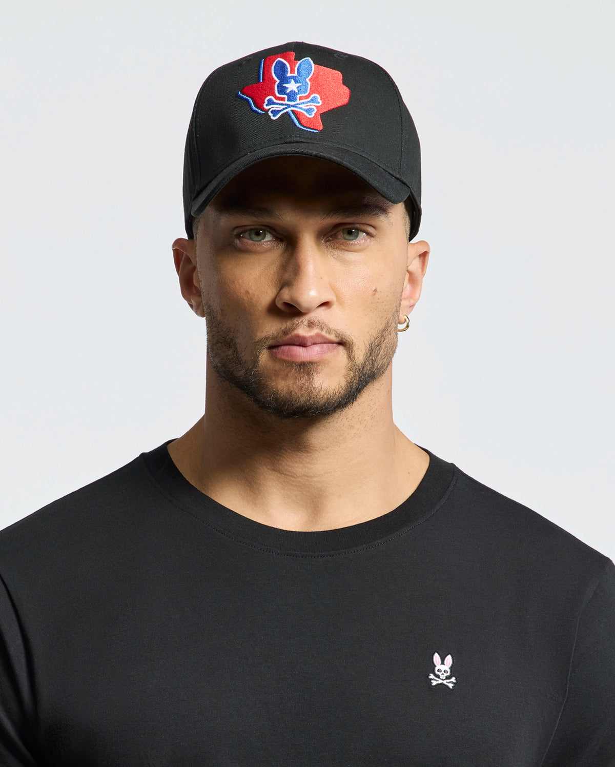 A man with light stubble wears a black Psycho Bunny MENS TEXAS BASEBALL CAP - MO1500014, featuring an embroidered red and blue skull design. He also sports a black shirt with a small woven skull and bunny logo against a plain white background.