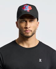 A man with light stubble wears a black Psycho Bunny MENS TEXAS BASEBALL CAP - MO1500014, featuring an embroidered red and blue skull design. He also sports a black shirt with a small woven skull and bunny logo against a plain white background.