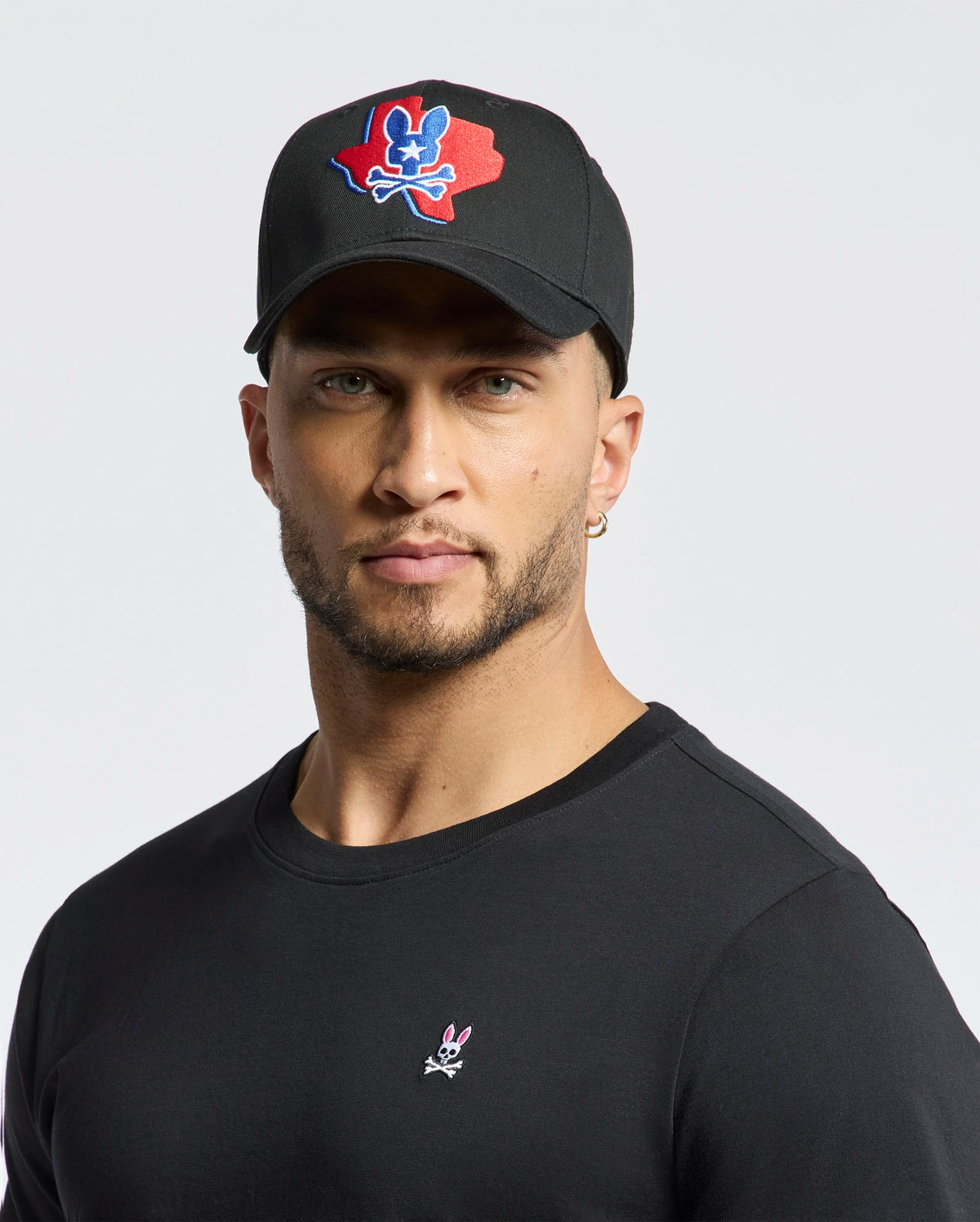 A man dons Psycho Bunny's MENS TEXAS BASEBALL CAP - MO1500014 with a colorful logo and a black cotton shirt embellished with a small design, set against a plain white background.